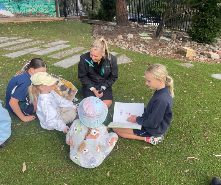 An SLSO sits outdoors with a group of young students as they read a picture book in the sunshine