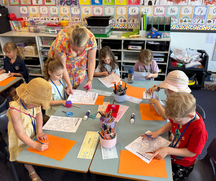 A teacher in a colourful dress helps new Kindergarten students to cut and paste a craft monkey